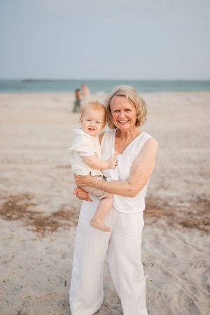 Smiling grandmother in white outfit holds happy baby on sunny beach with ocean in background.