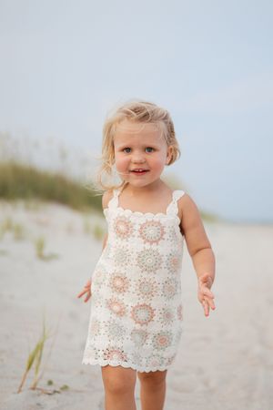 Smiling toddler girl in white crochet dress playing on sandy beach with dunes in background.