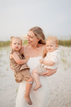 A smiling woman in a white dress holds two toddlers on a sandy beach, with dunes and soft sky in the background.