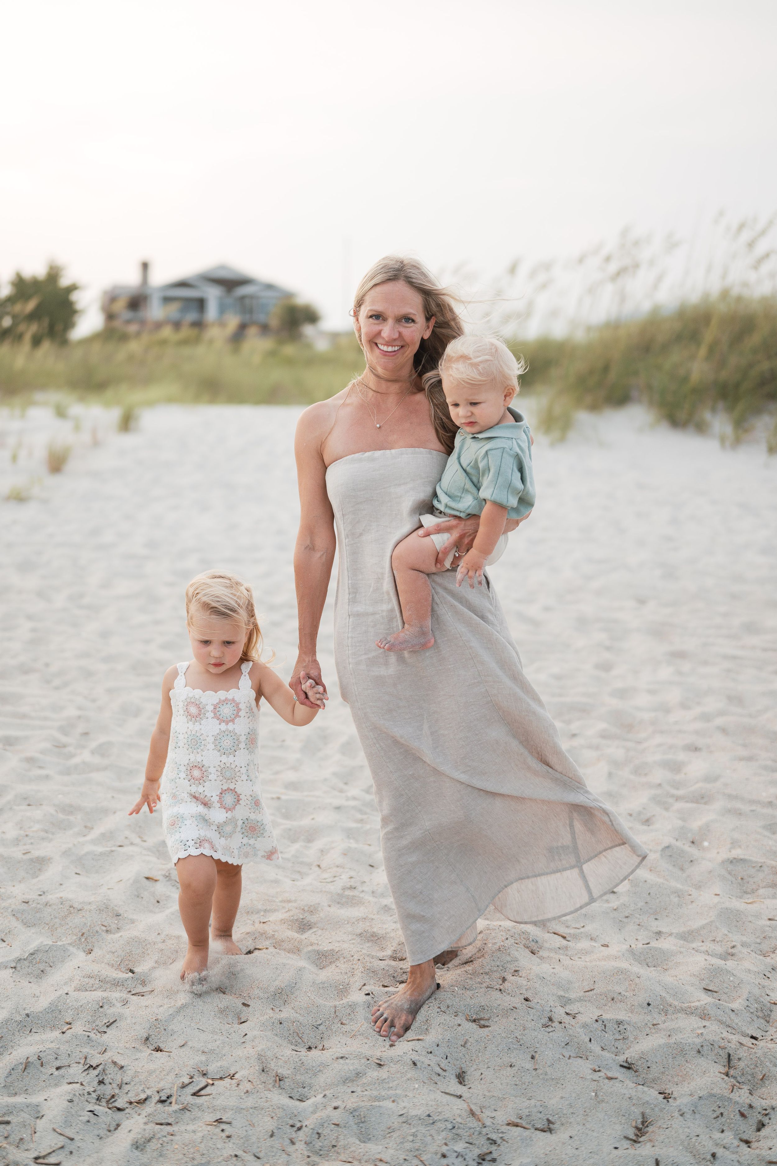 Smiling mother in gray maxi dress holds toddler son while walking hand-in-hand with young daughter on beach.