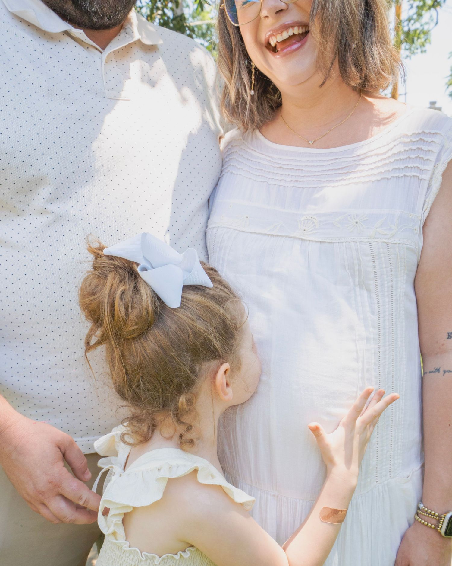 Pregnant woman in white dress laughing as young daughter touches her baby bump outdoors.