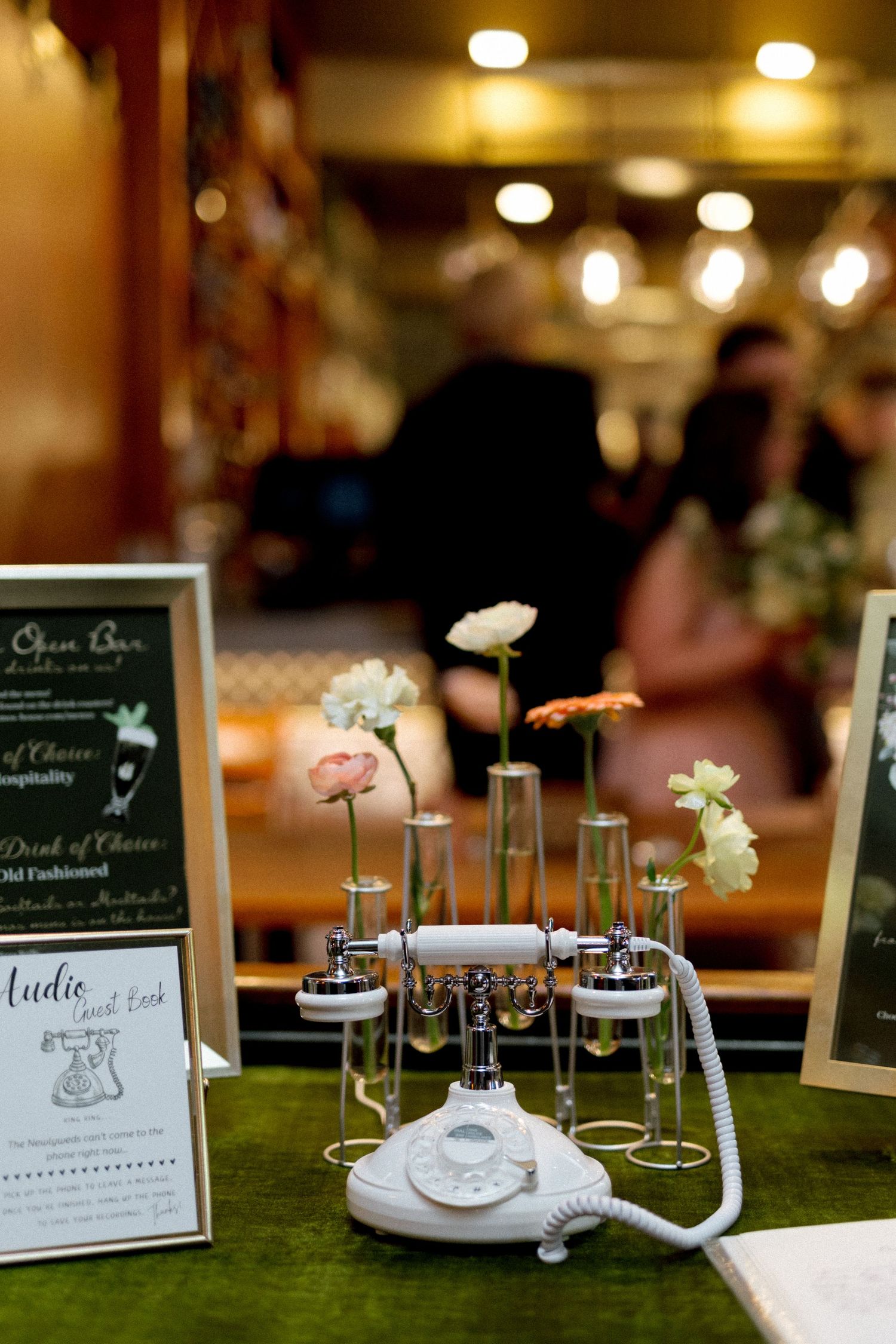 Vintage white rotary phone with bud vases holding pink flowers on a green velvet table at a wedding reception.