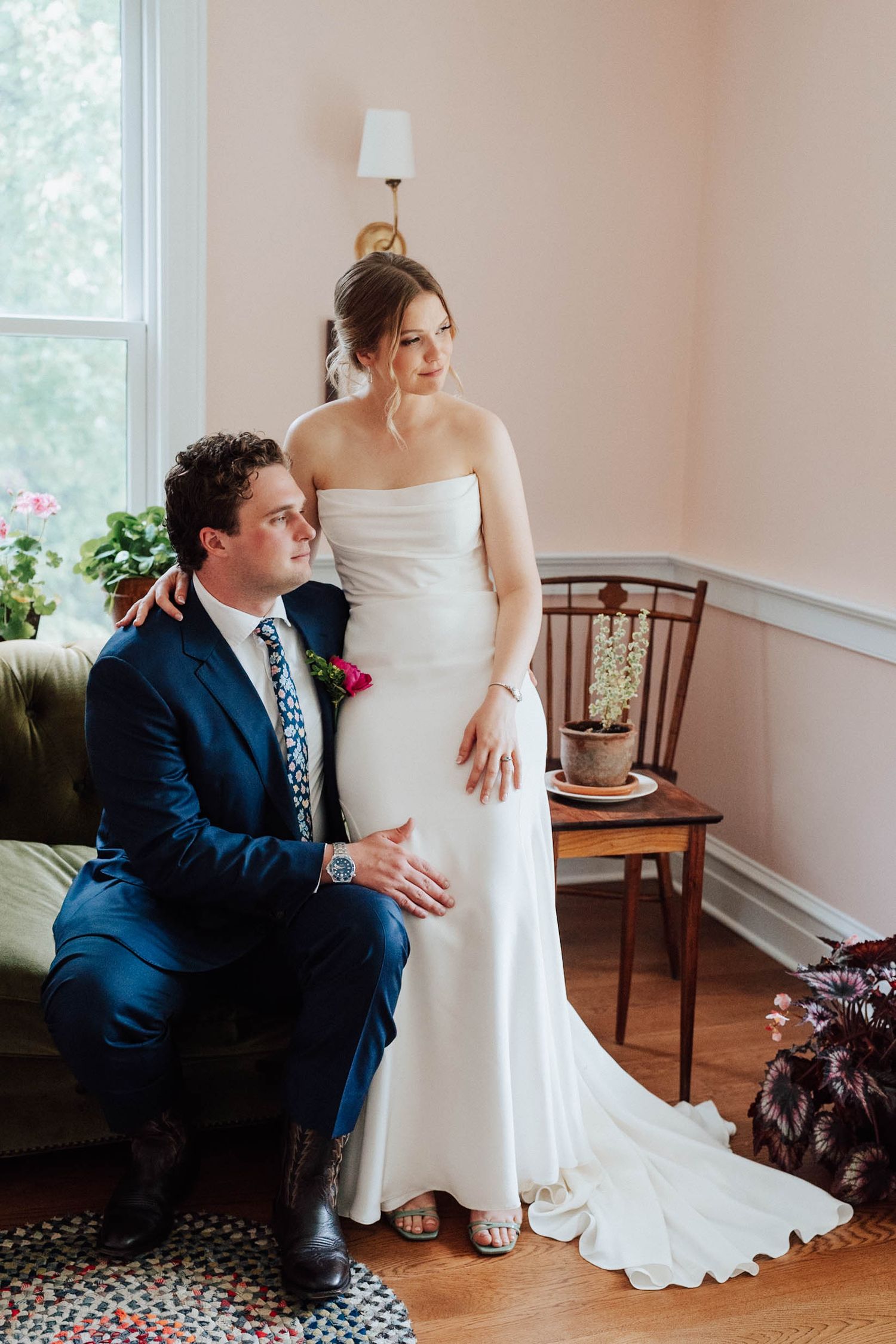 Bride in white strapless gown stands beside groom in navy suit seated on green sofa in elegant room.