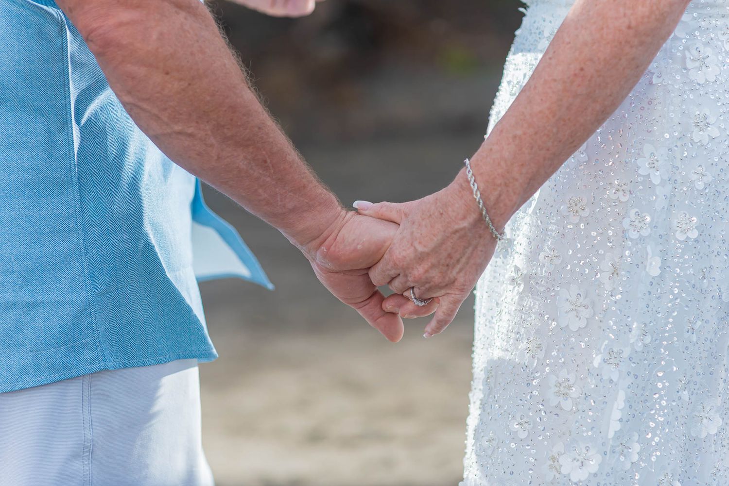 Elderly couple holding hands at beach wedding, bride in white lace dress and groom in light blue shirt.