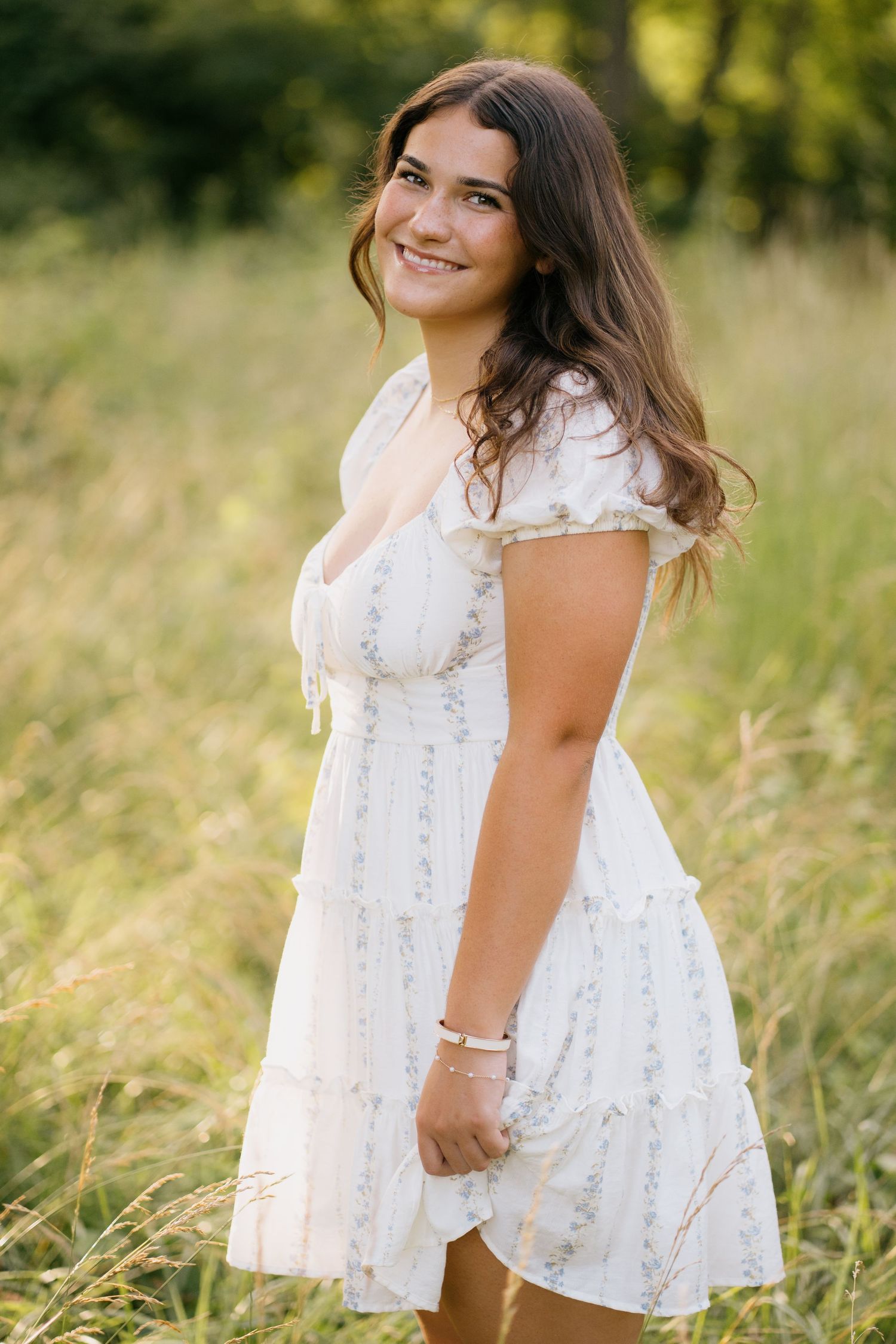 Young woman in white floral dress smiling in a sunlit meadow with tall grass and trees in background.