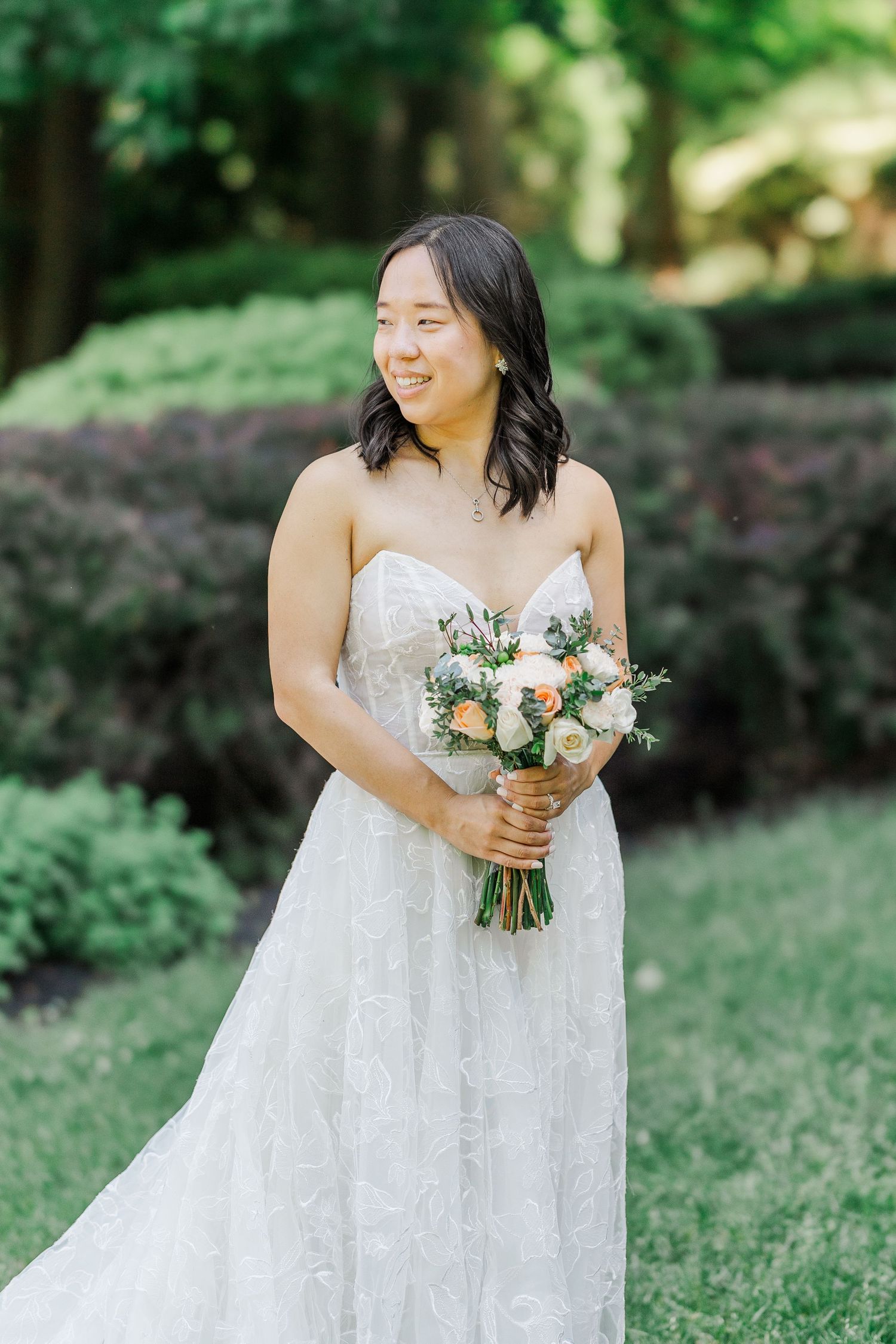 Smiling bride in strapless lace wedding gown holds peach and white bouquet outdoors in lush green garden setting.