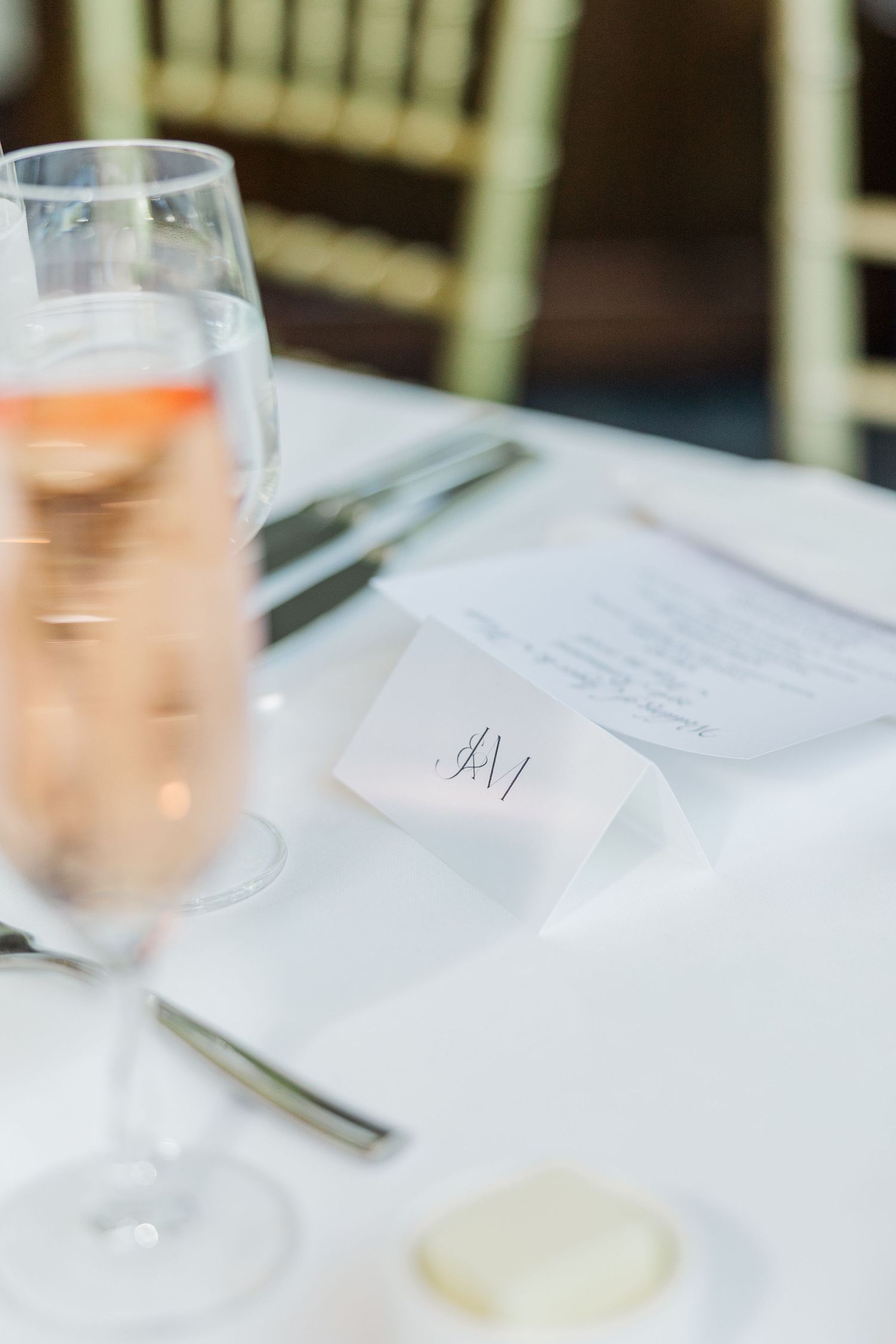 Elegant wedding reception table with rose champagne, monogram place card, menu, and cutlery on white linen.