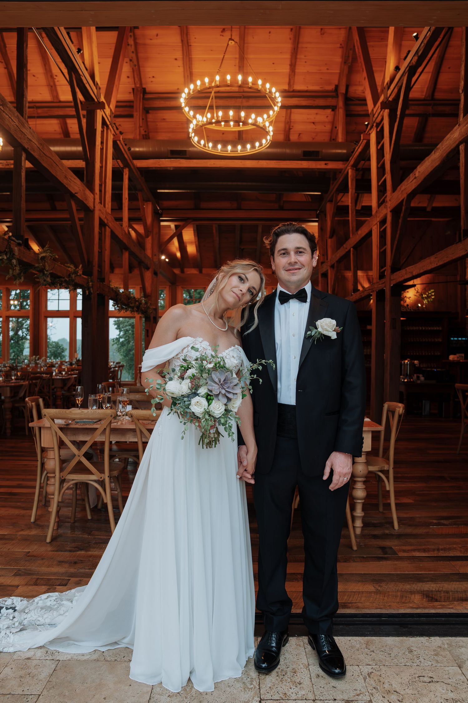 Bride and groom pose together inside a warmly lit rustic barn venue holding a floral bouquet.