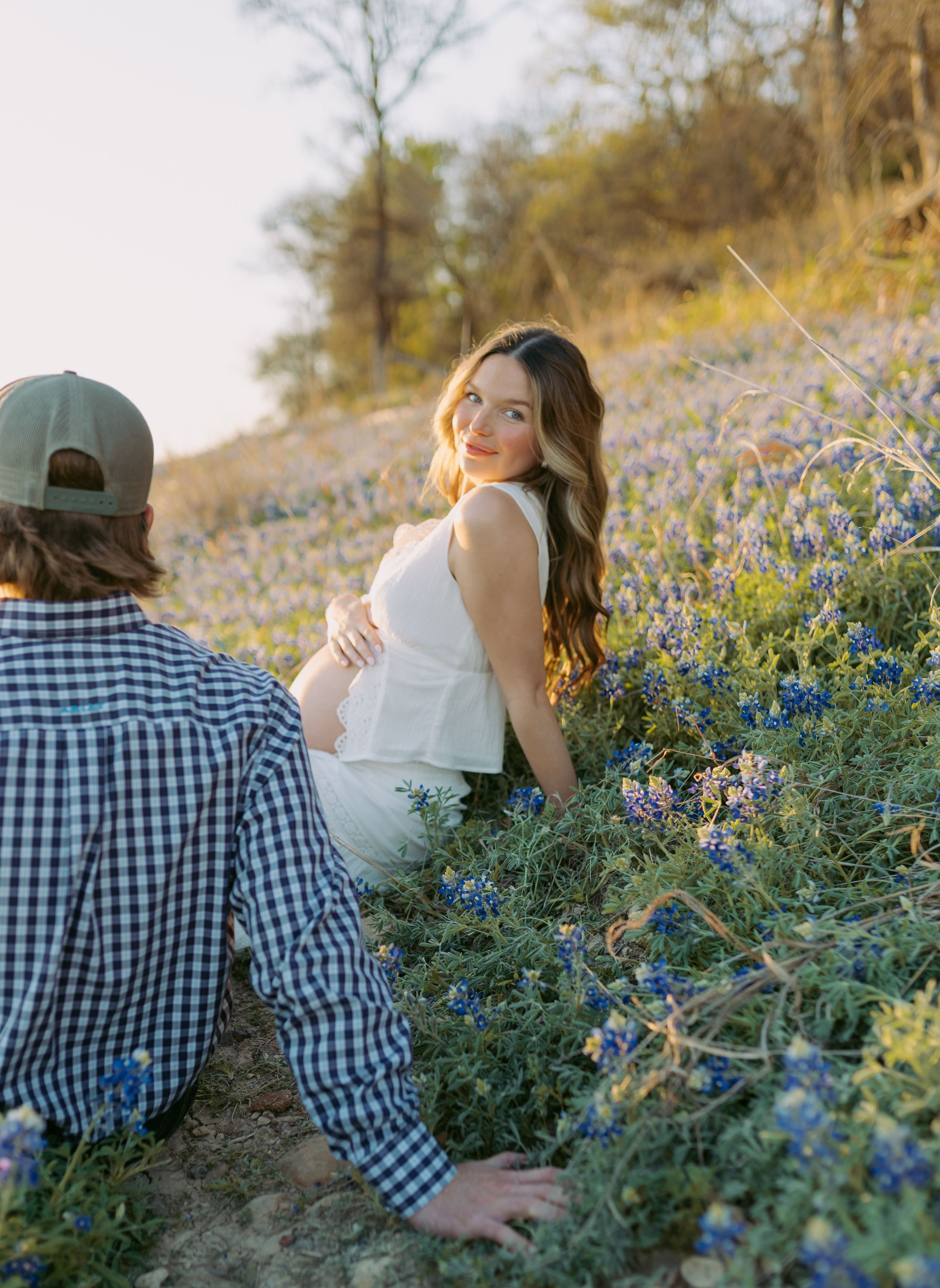 A couple sits among blooming bluebonnets on a hillside during golden hour in spring.