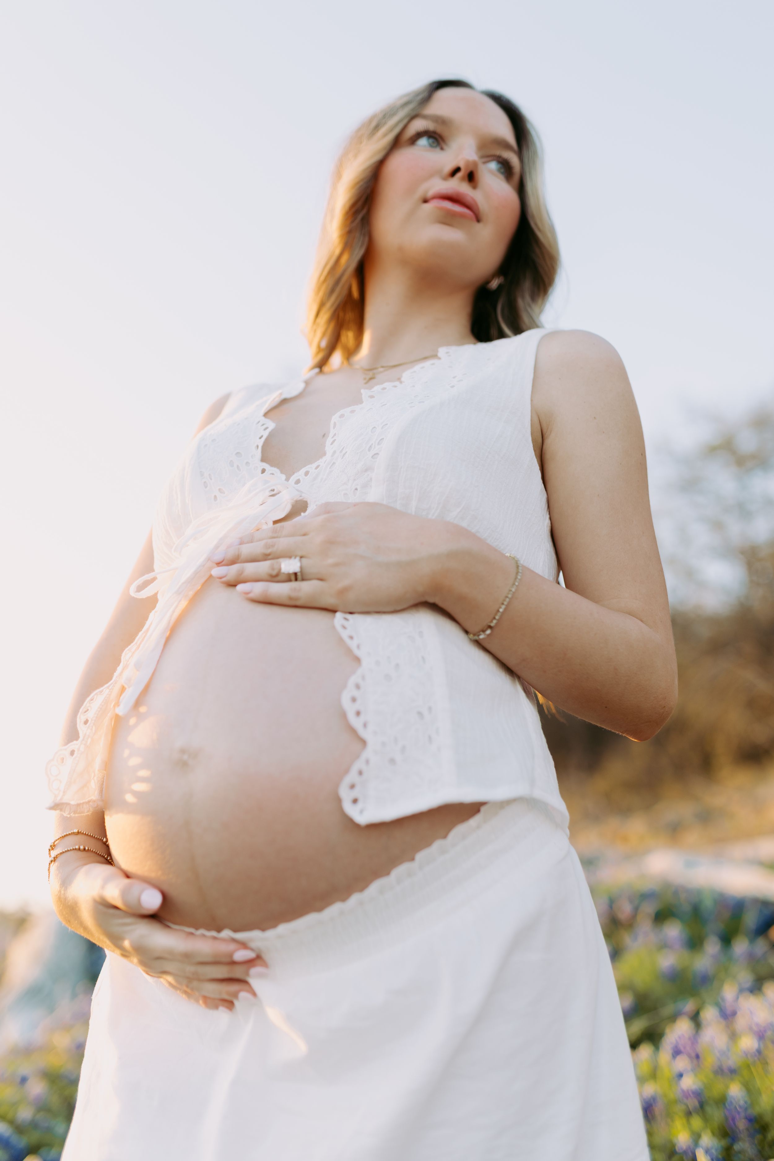 A pregnant woman in a white dress cradles her baby bump while standing in a field of wildflowers at sunset.