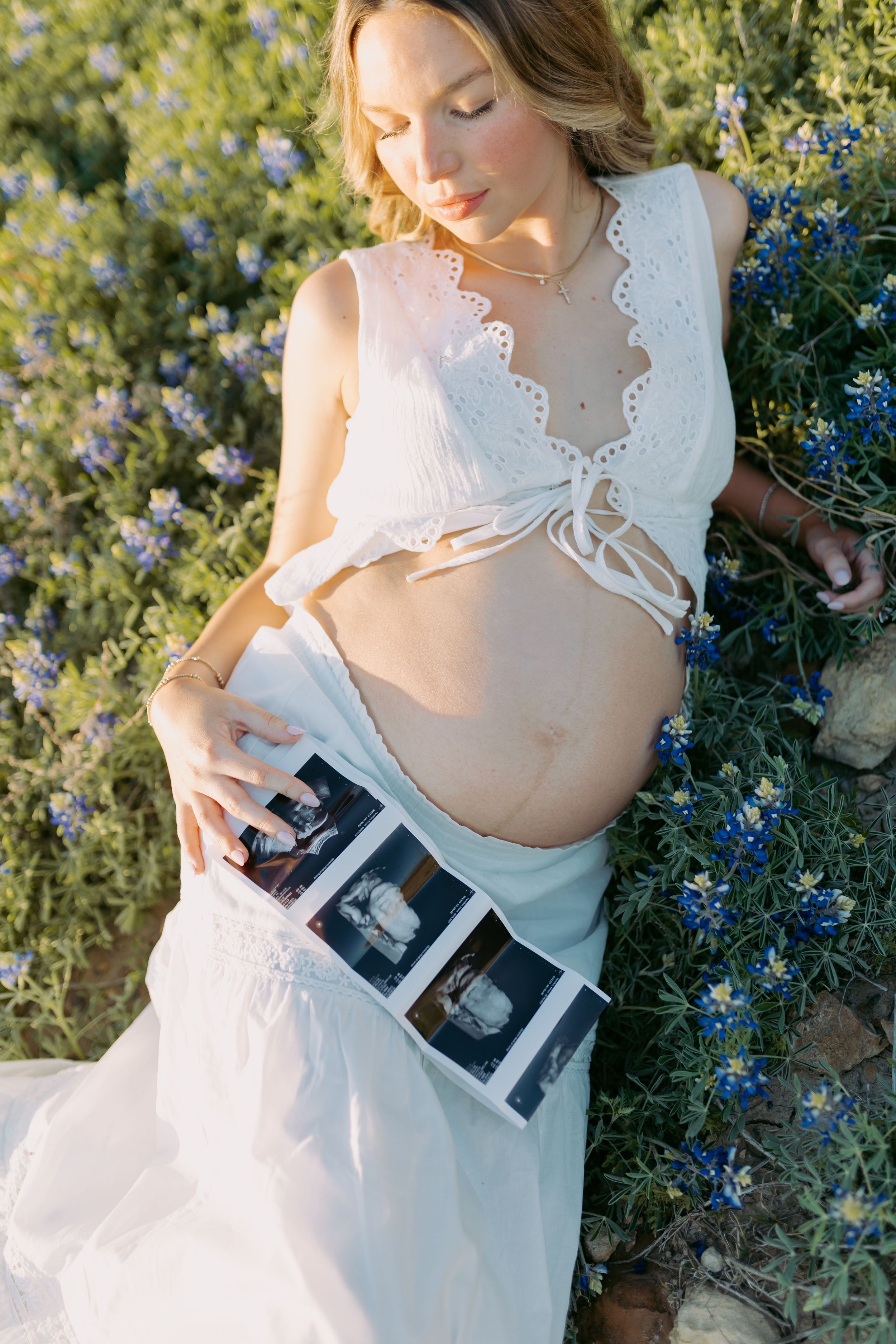 A person in a white dress holds ultrasound photos while posing in a garden with blue flowers.