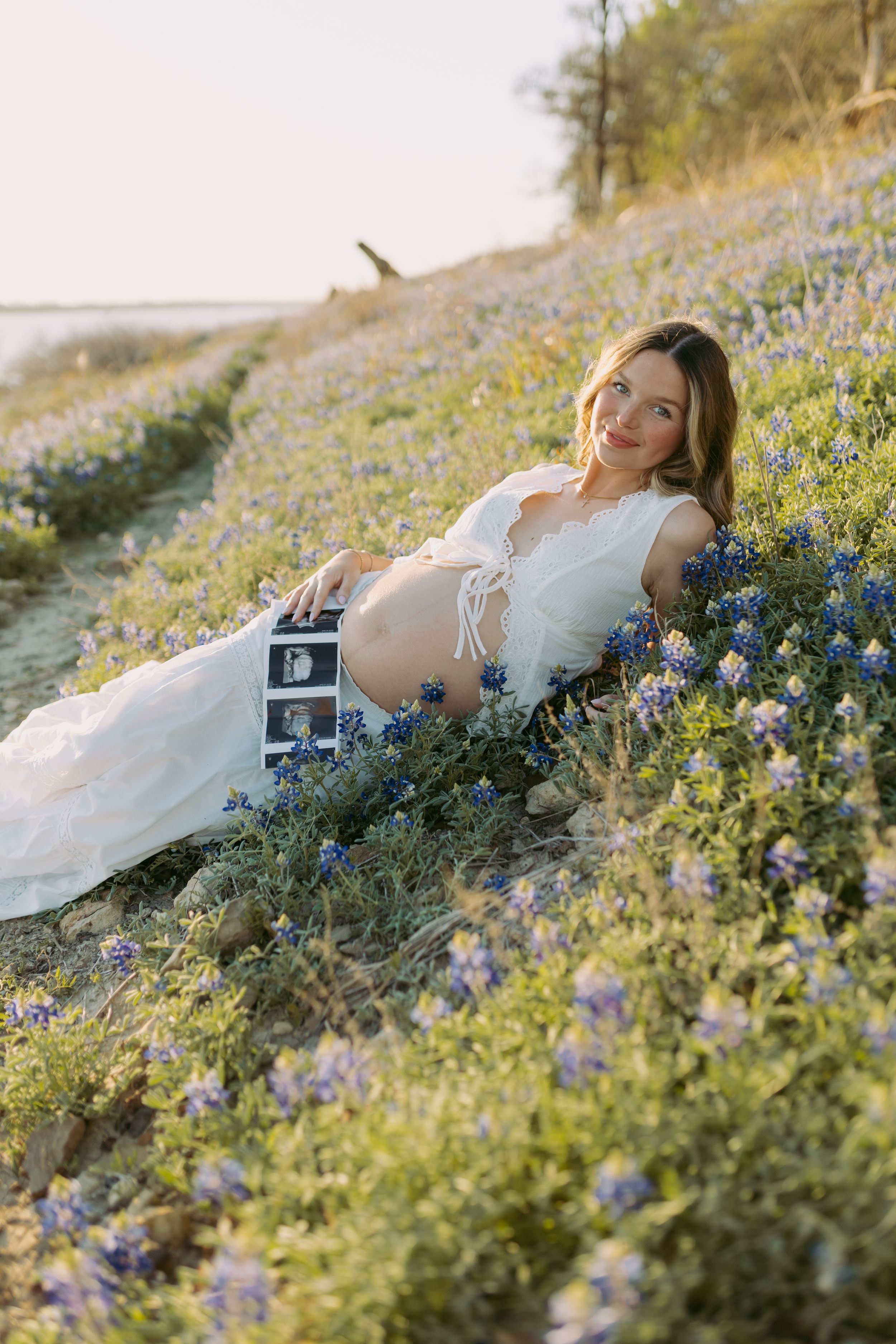 A maternity photo session in a field of blooming bluebonnets at sunset with a sonogram photo displayed.