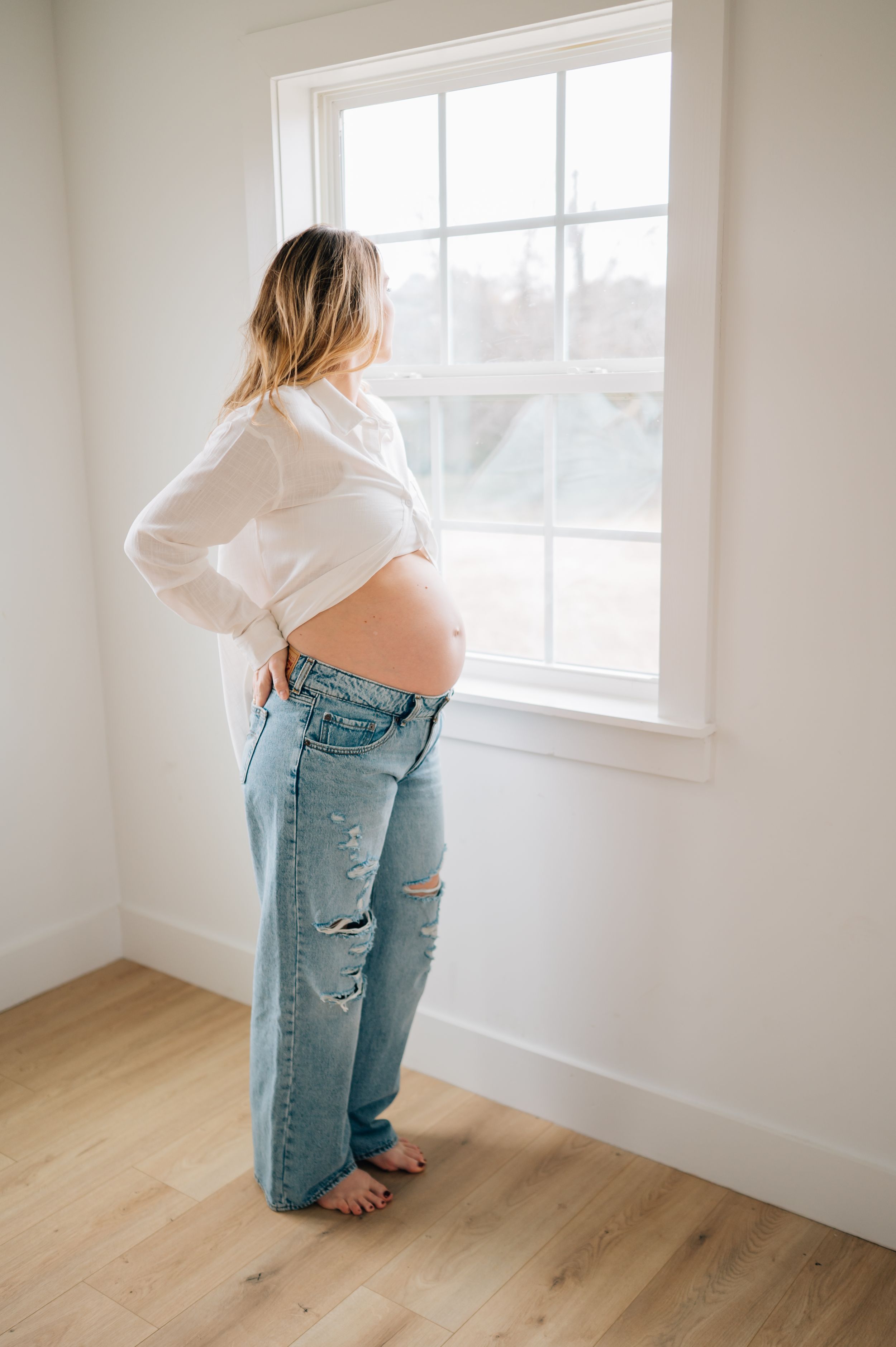 Woman in white top and distressed jeans sits near bright window in empty room with hardwood floors.