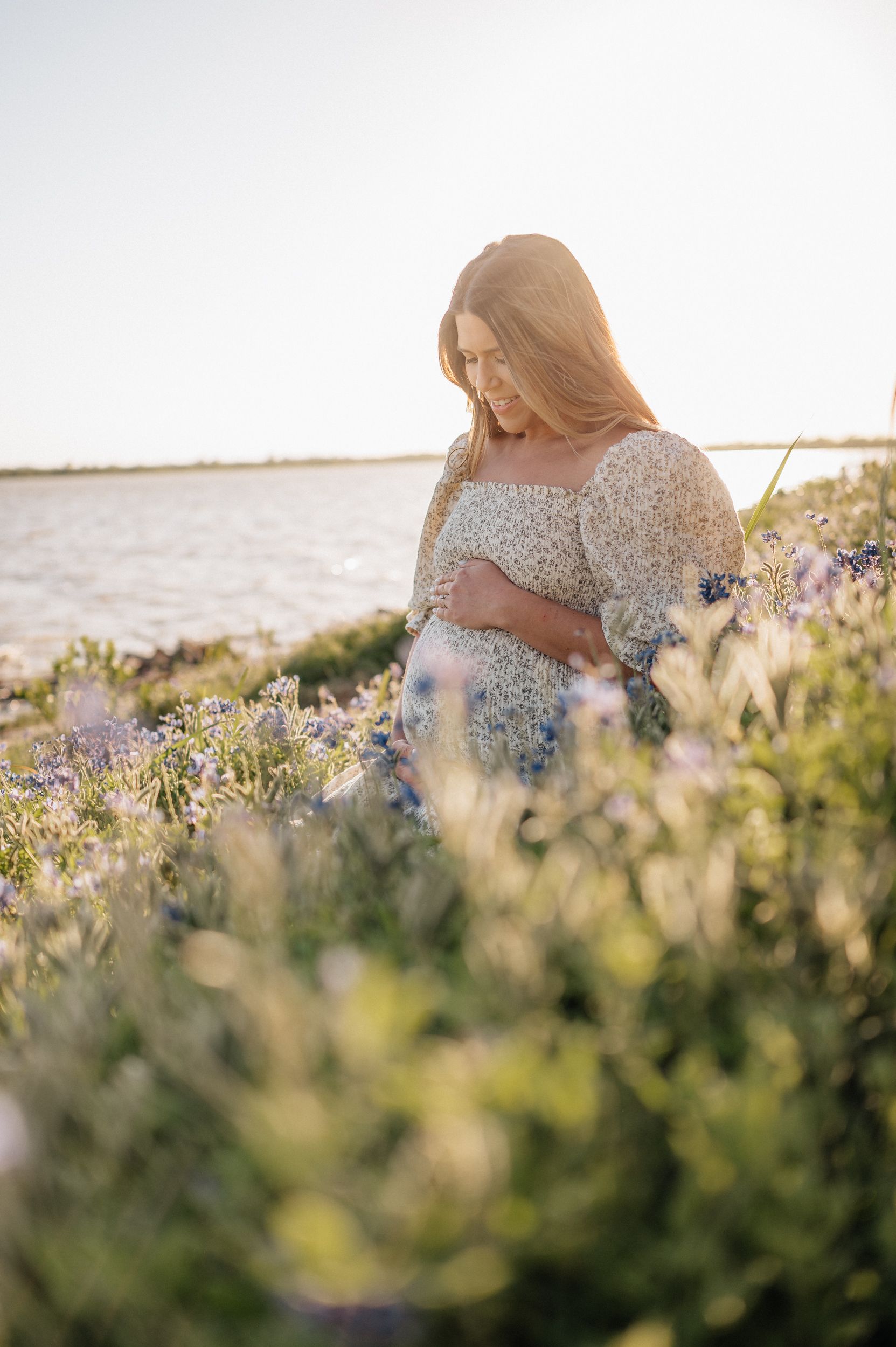 Woman in floral dress standing amid wildflowers at sunset near waterfront.