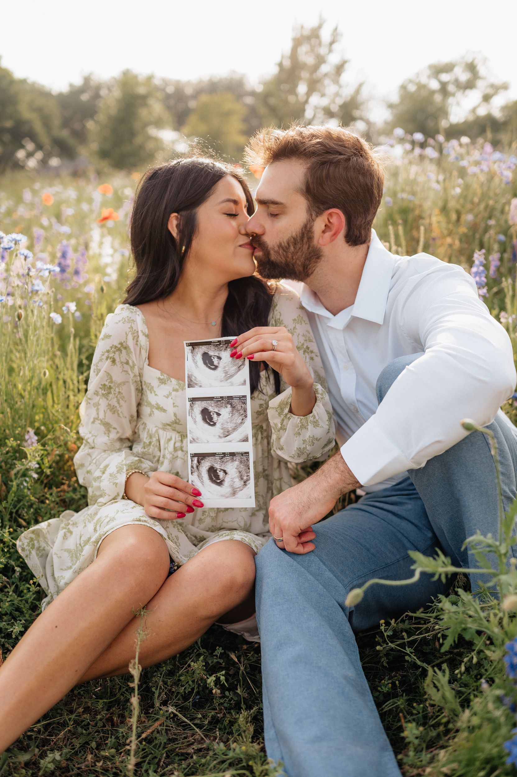 Couple sharing intimate moment in flower field while holding ultrasound photos.