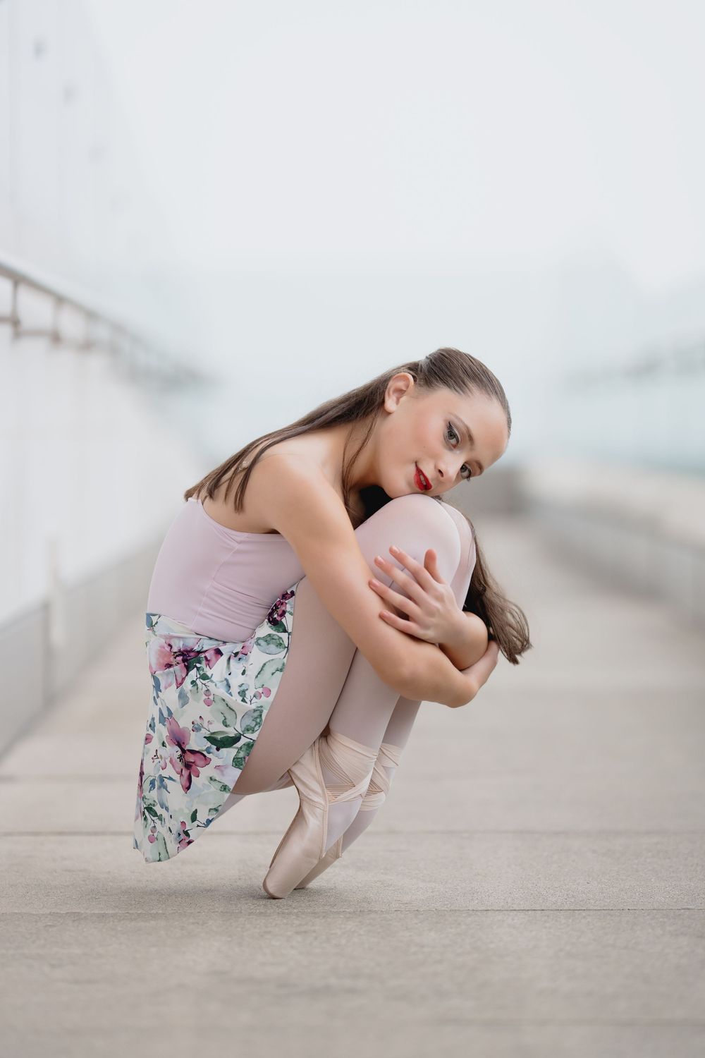 Ballet dancer in floral skirt and pink leotard performs graceful poses on light colored floor.