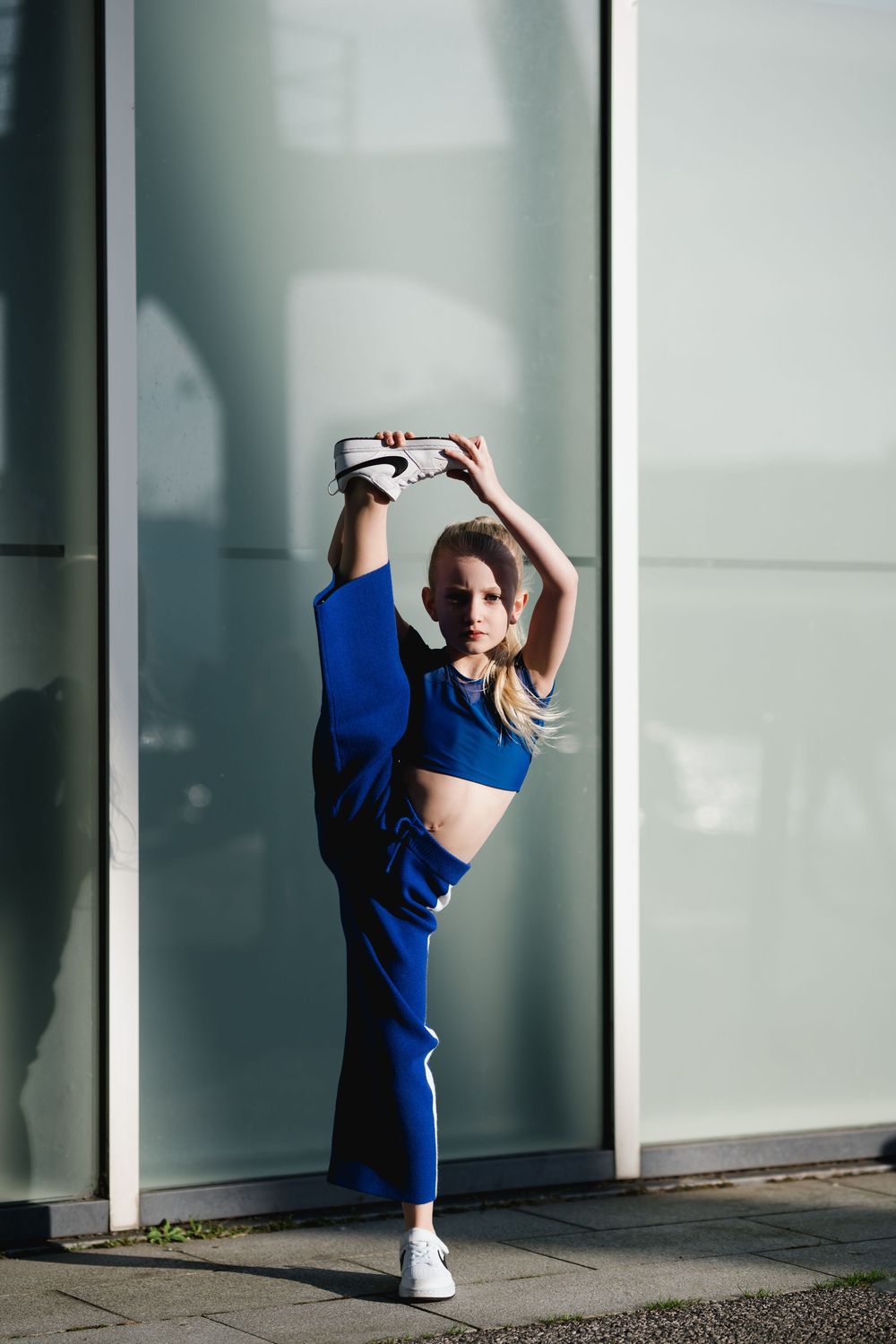 A woman in a blue outfit performs stretching exercises against a white wall with dramatic shadows.