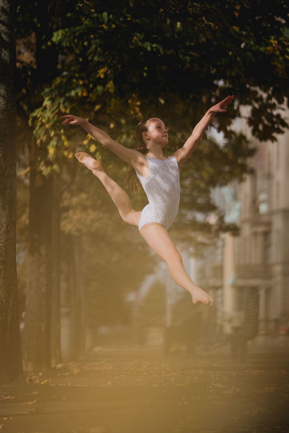 Graceful ballet dancer leaps through autumn air in white leotard as golden sunlight filters through trees.