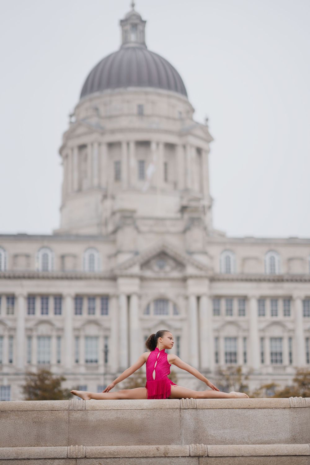 A gymnast in a pink leotard performs a split pose in front of the Port of Liverpool Building's domed architecture.