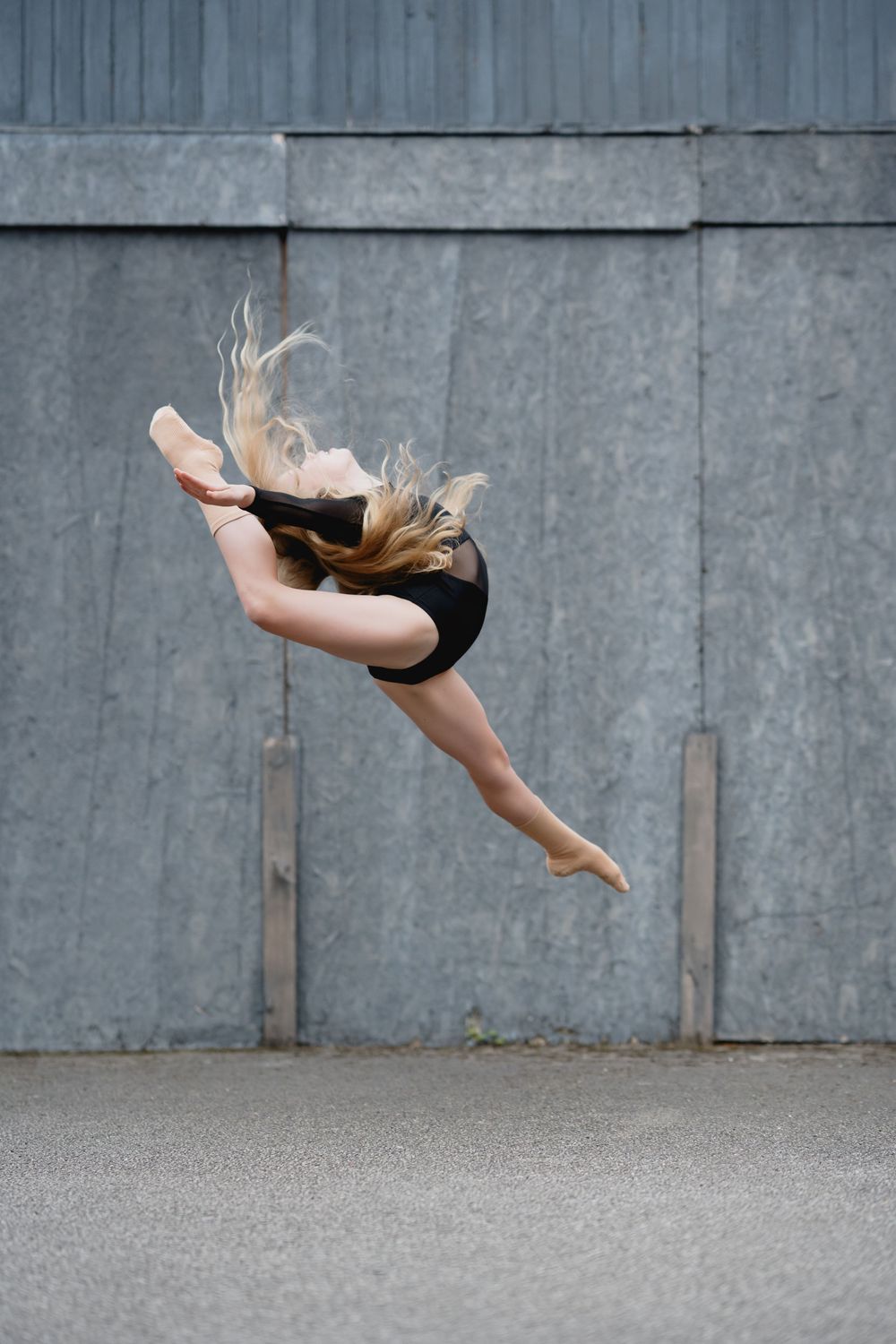 A dancer executes a dynamic horizontal leap against an industrial concrete wall backdrop.
