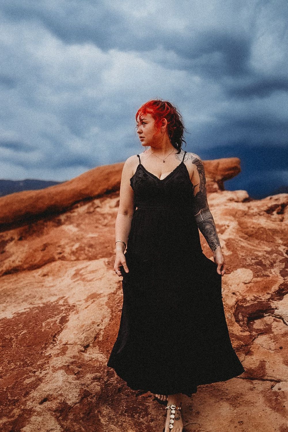 Woman with red hair in black dress standing on red rocks under dramatic stormy sky.