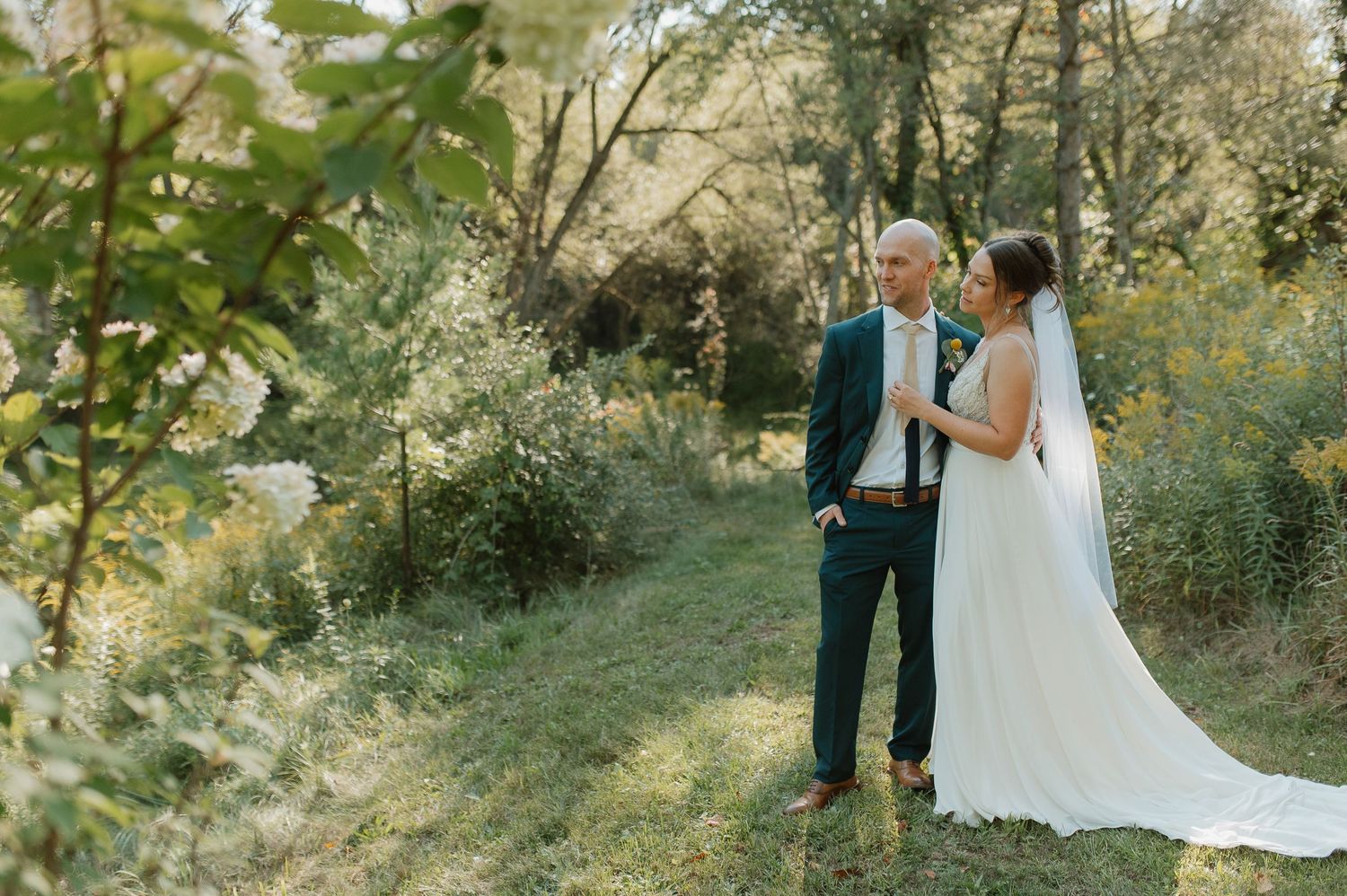 A couple in wedding attire standing together in a lush green outdoor setting.