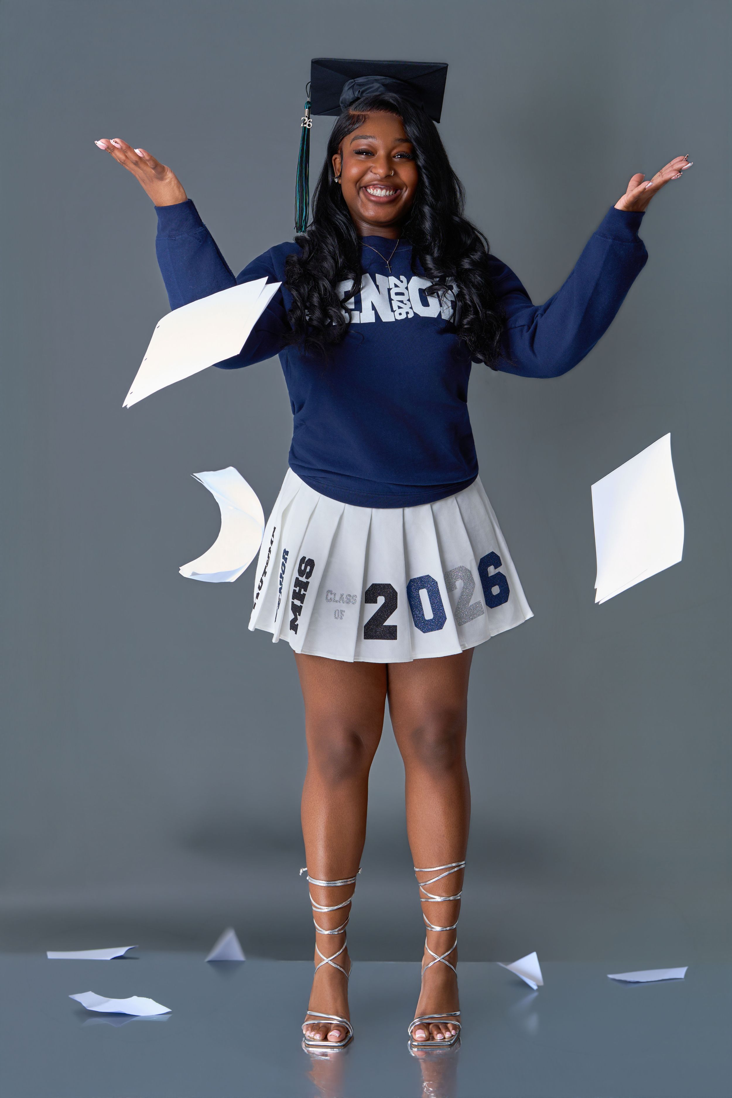 Smiling graduate in cap and senior sweatshirt with Class of 2026 skirt tosses papers in studio photo.