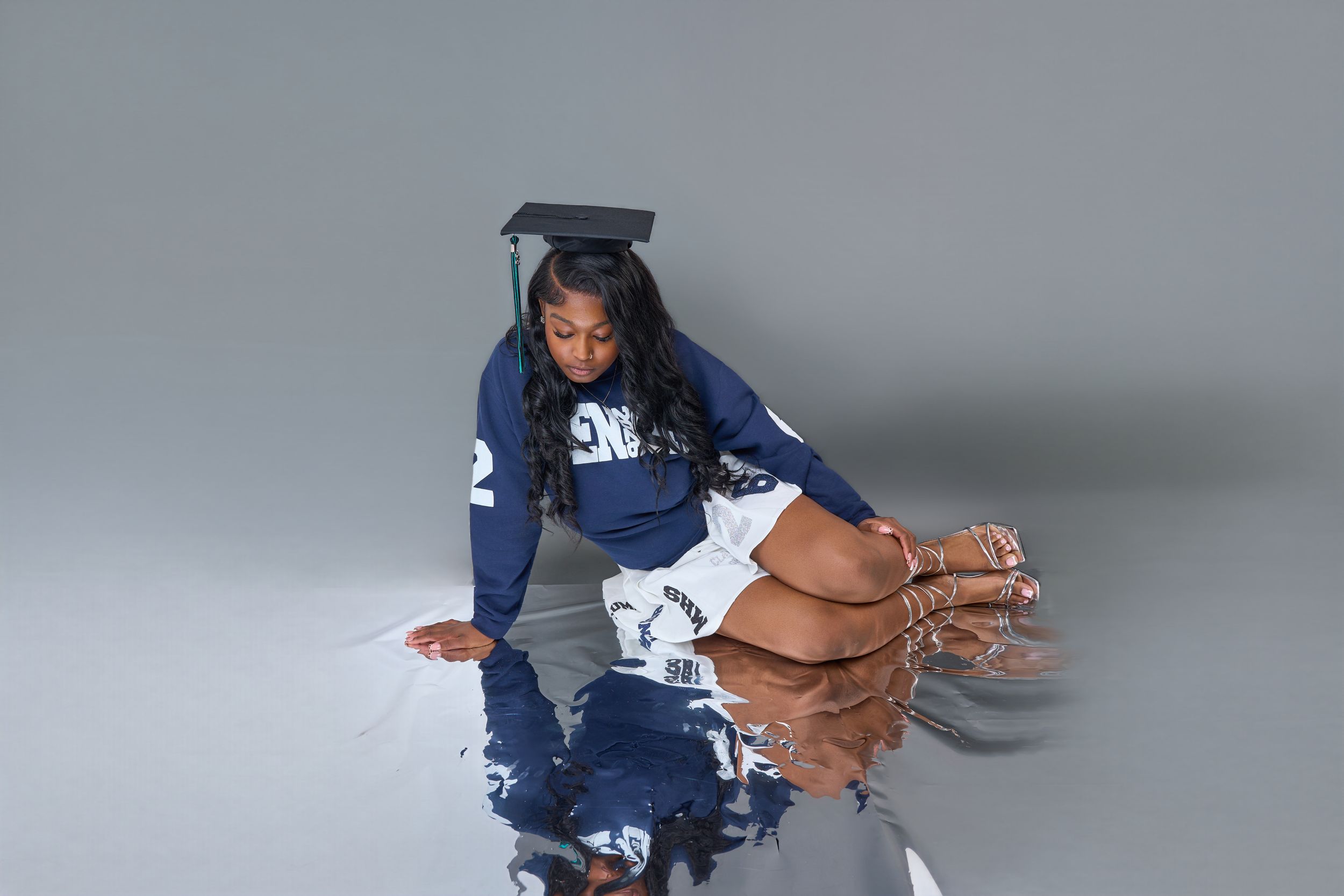 Young woman in graduation cap and blue jersey poses on reflective floor in professional grad photo.