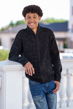Smiling teenage boy in dark knit zip sweater and jeans leaning on white railing outdoors.