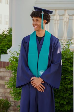 Smiling young man in navy blue graduation gown and teal stole posing outdoors near white columns and green shrubs.
