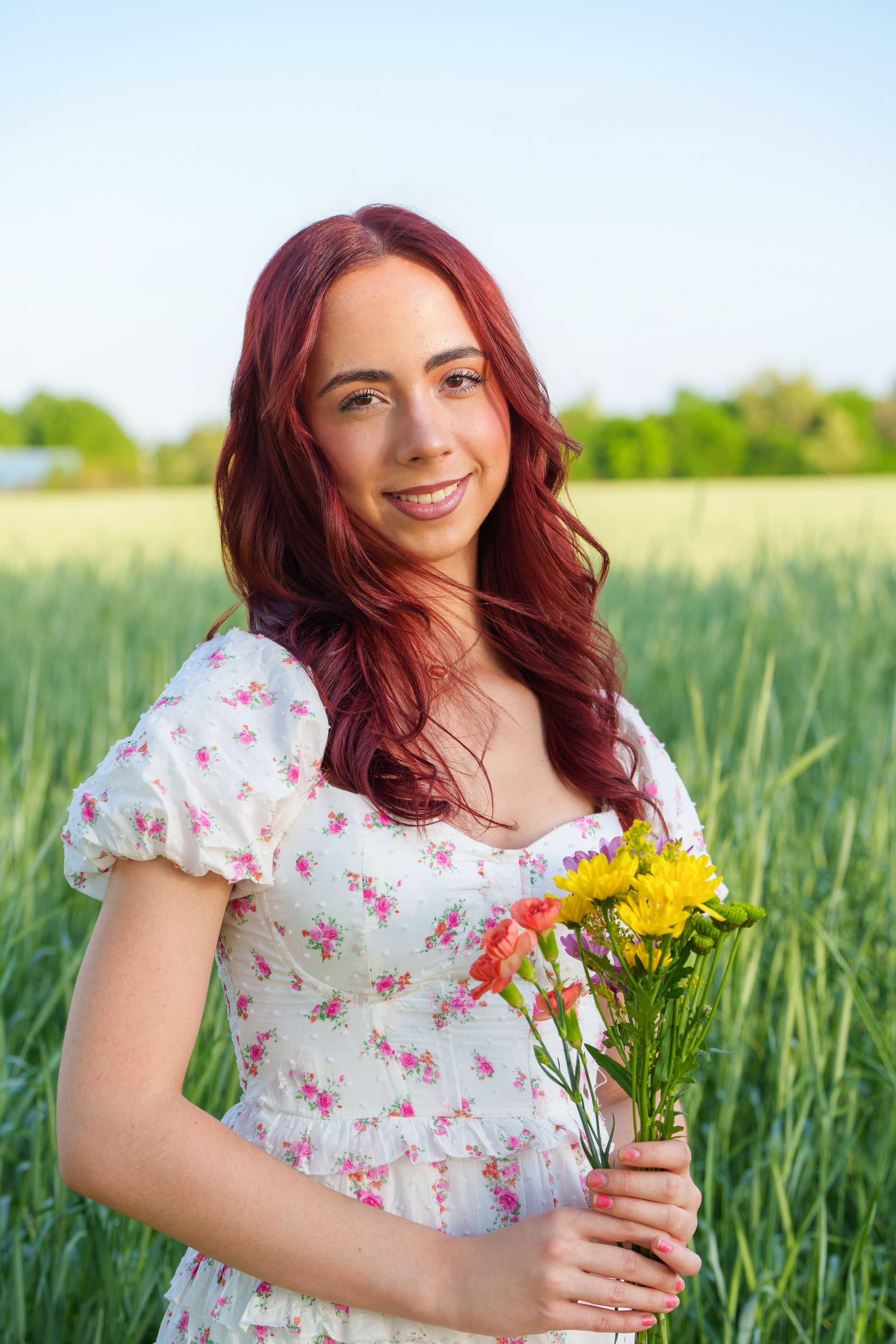 Red-haired woman in floral dress holding colorful flowers in a green field on a sunny day.