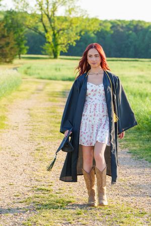 Young woman with red hair wearing graduation gown over floral dress and cowboy boots on a sunny rural path.