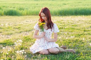 Young woman in floral dress sitting on grass, smelling yellow sunflowers in a sunny green meadow.
