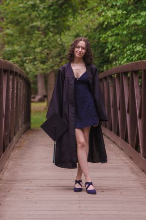 Young woman in graduation gown and cap posing on a wooden bridge surrounded by lush green trees.