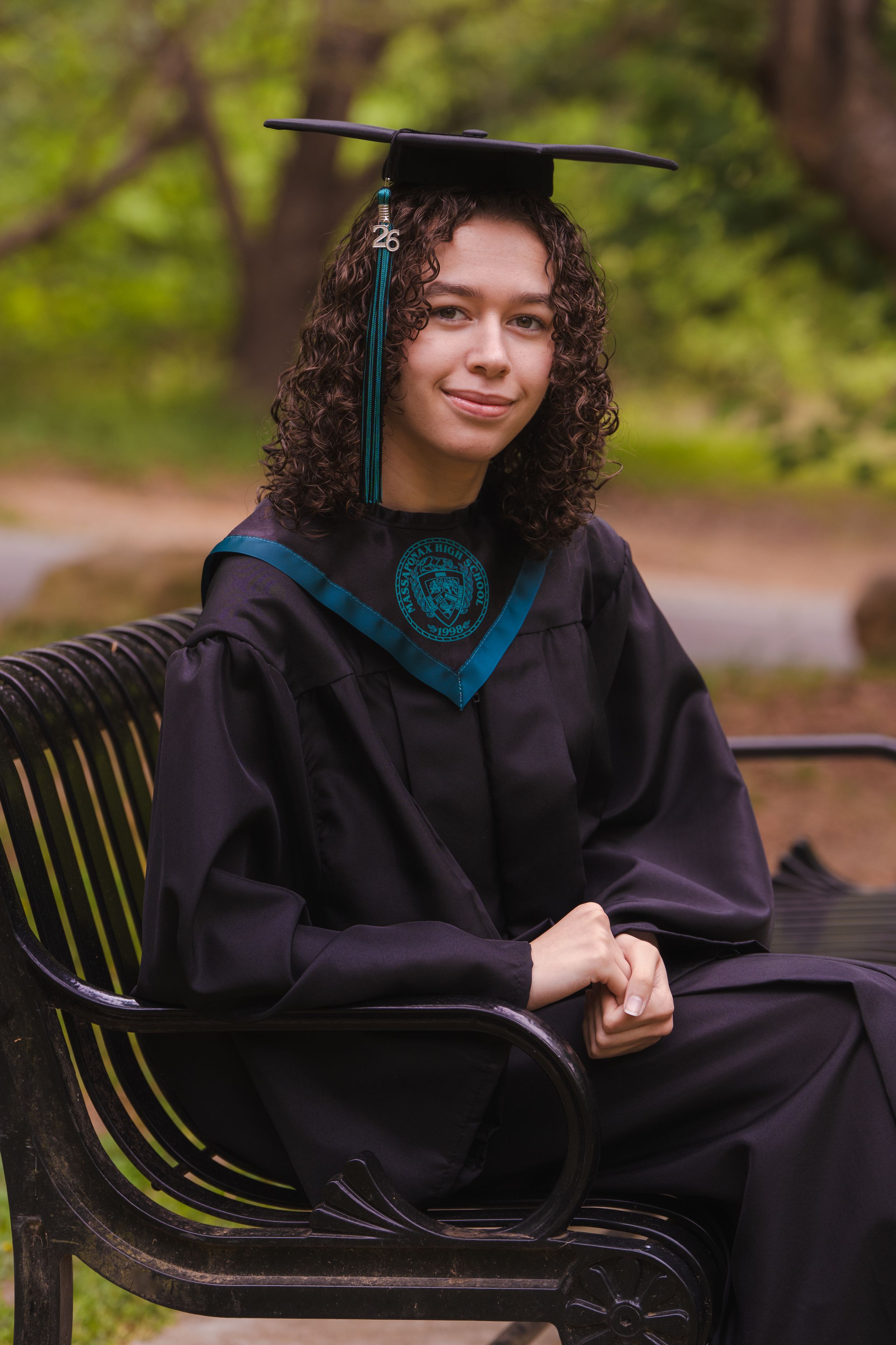 Young woman in black graduation gown and cap sits on bench outdoors, smiling with curly hair and teal academic regalia.