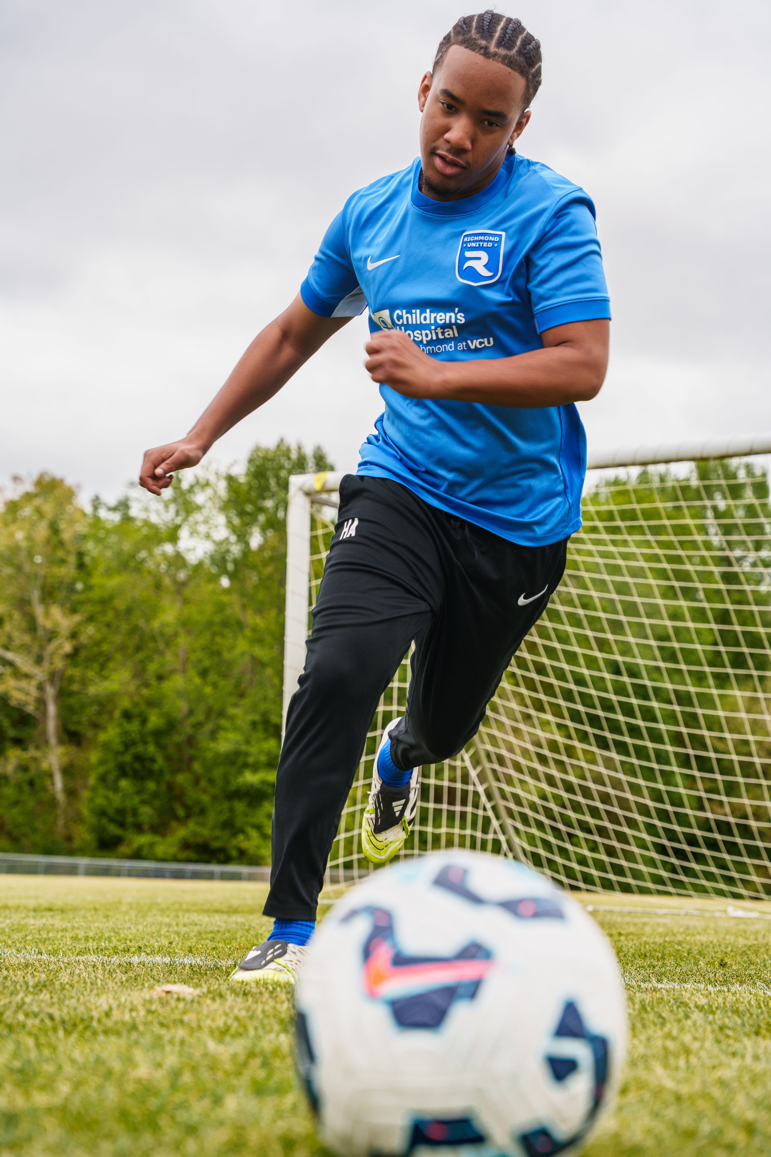 Young soccer player in blue Nike jersey kicking ball on green field with goal net in background.