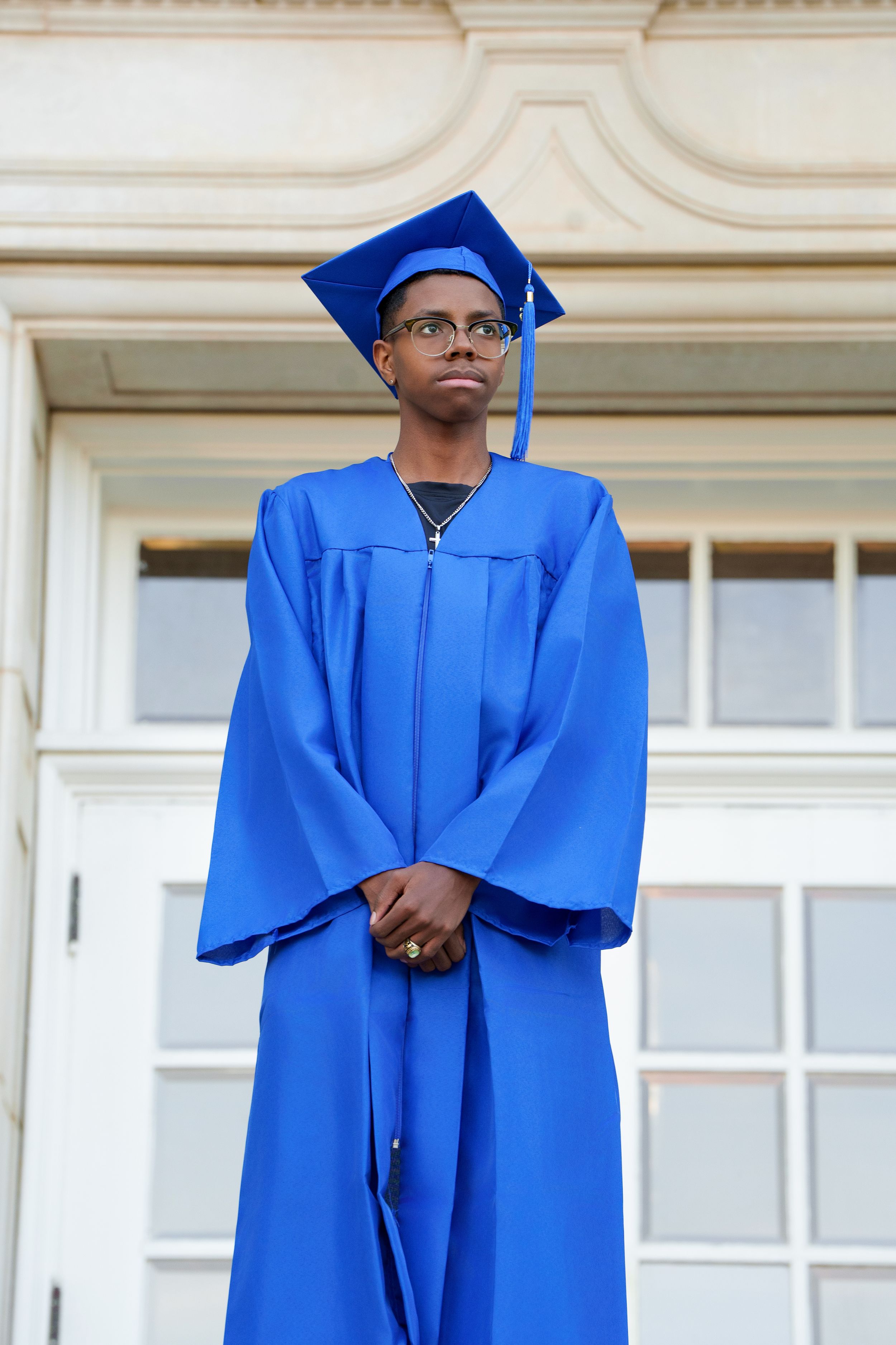 Young graduate in blue cap and gown stands proudly before a classical building, gazing upward with quiet confidence.