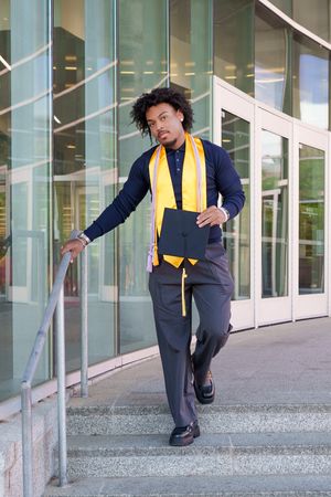 Young Black male graduate in navy suit with gold sash holds cap on building steps, celebrating academic achievement.