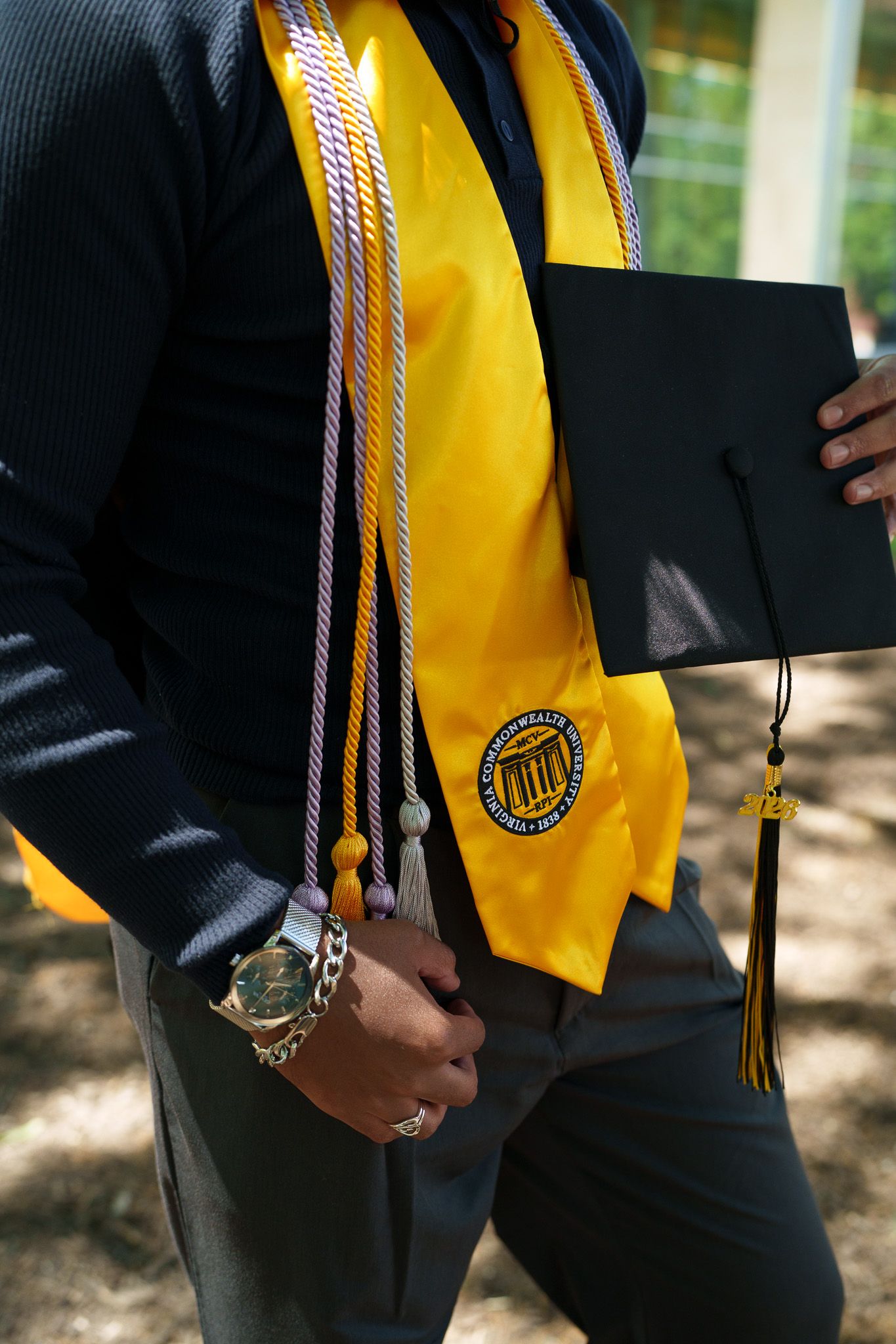 Graduate wearing gold stole with honor cords holding black mortarboard cap at outdoor graduation ceremony.