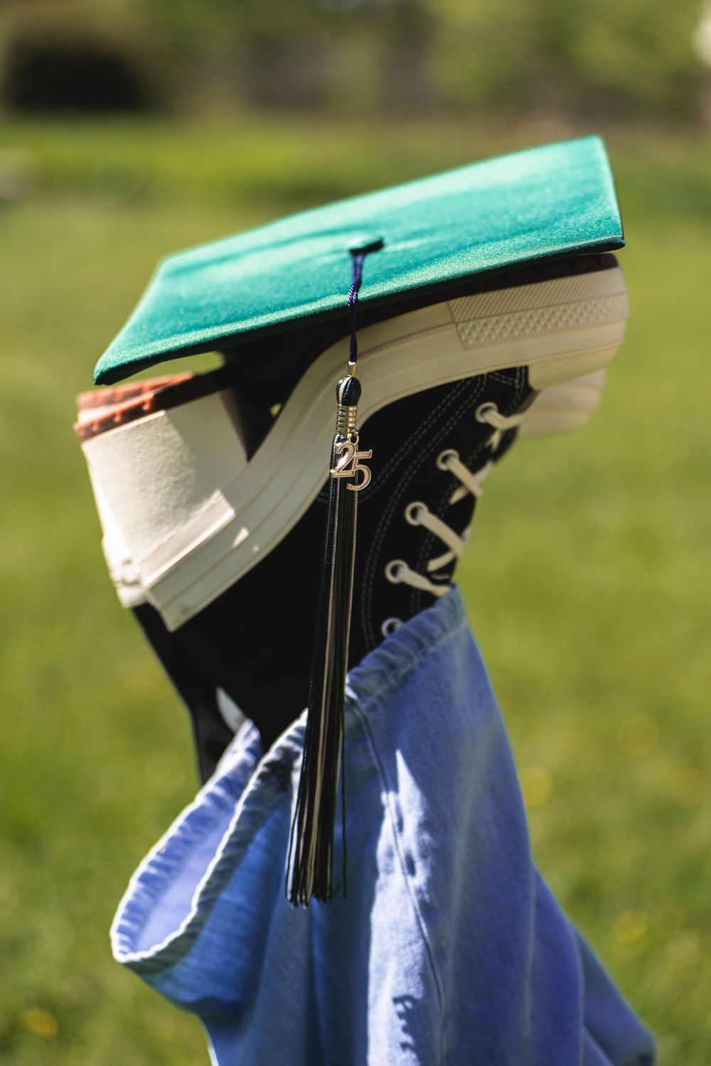 Close-up of graduation cap with tassel and black sneakers against green background.