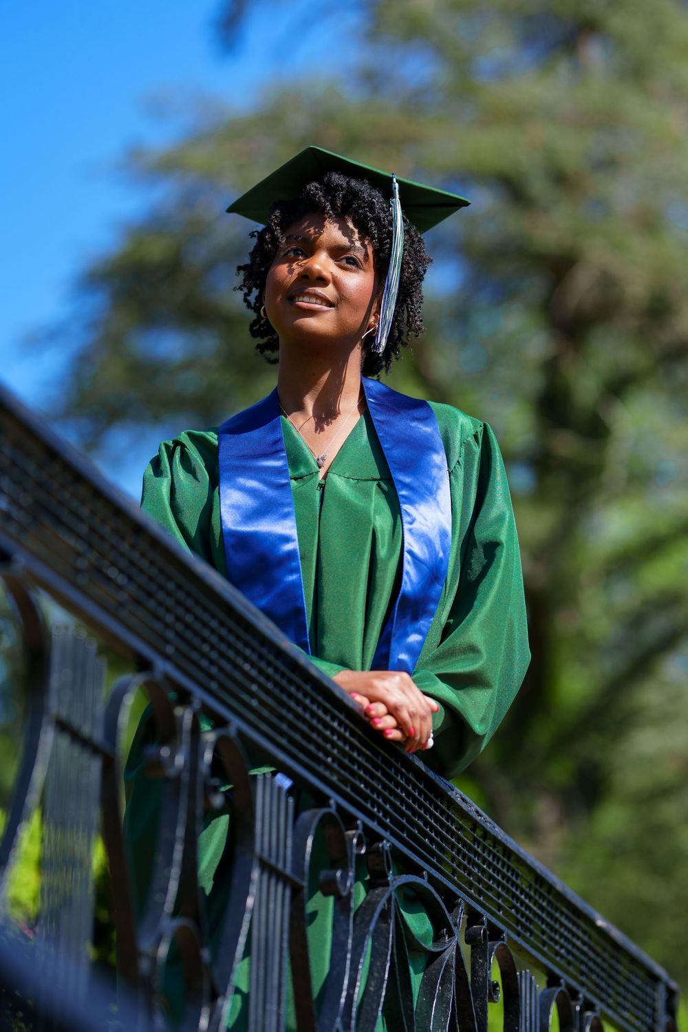 Graduate wearing green gown and blue stole standing on outdoor staircase railing.