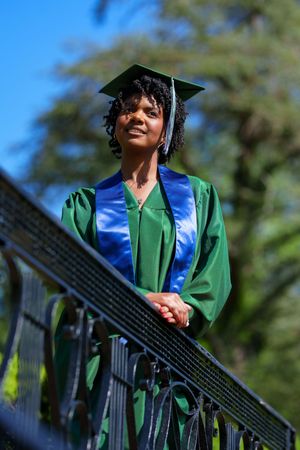 Graduate wearing green gown and blue stole standing on outdoor staircase railing.