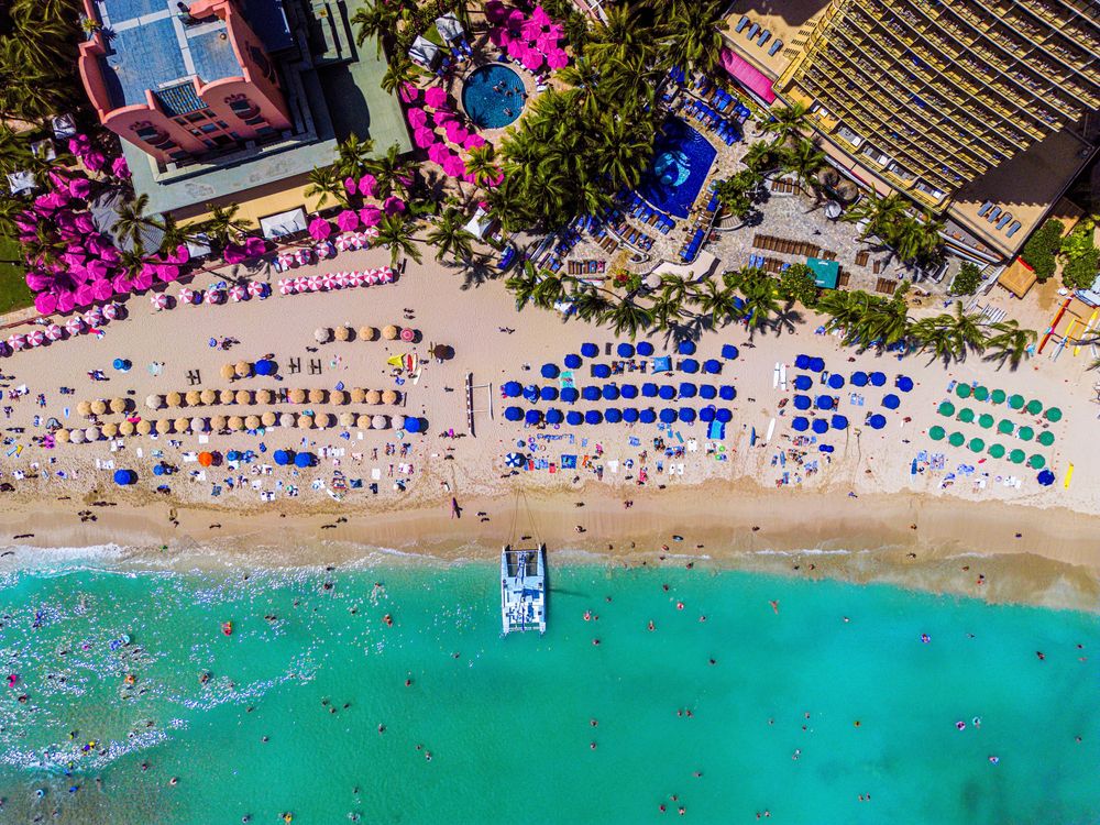Aerial view of colorful beach resort with blue umbrellas, turquoise water, and crowded sandy beach.