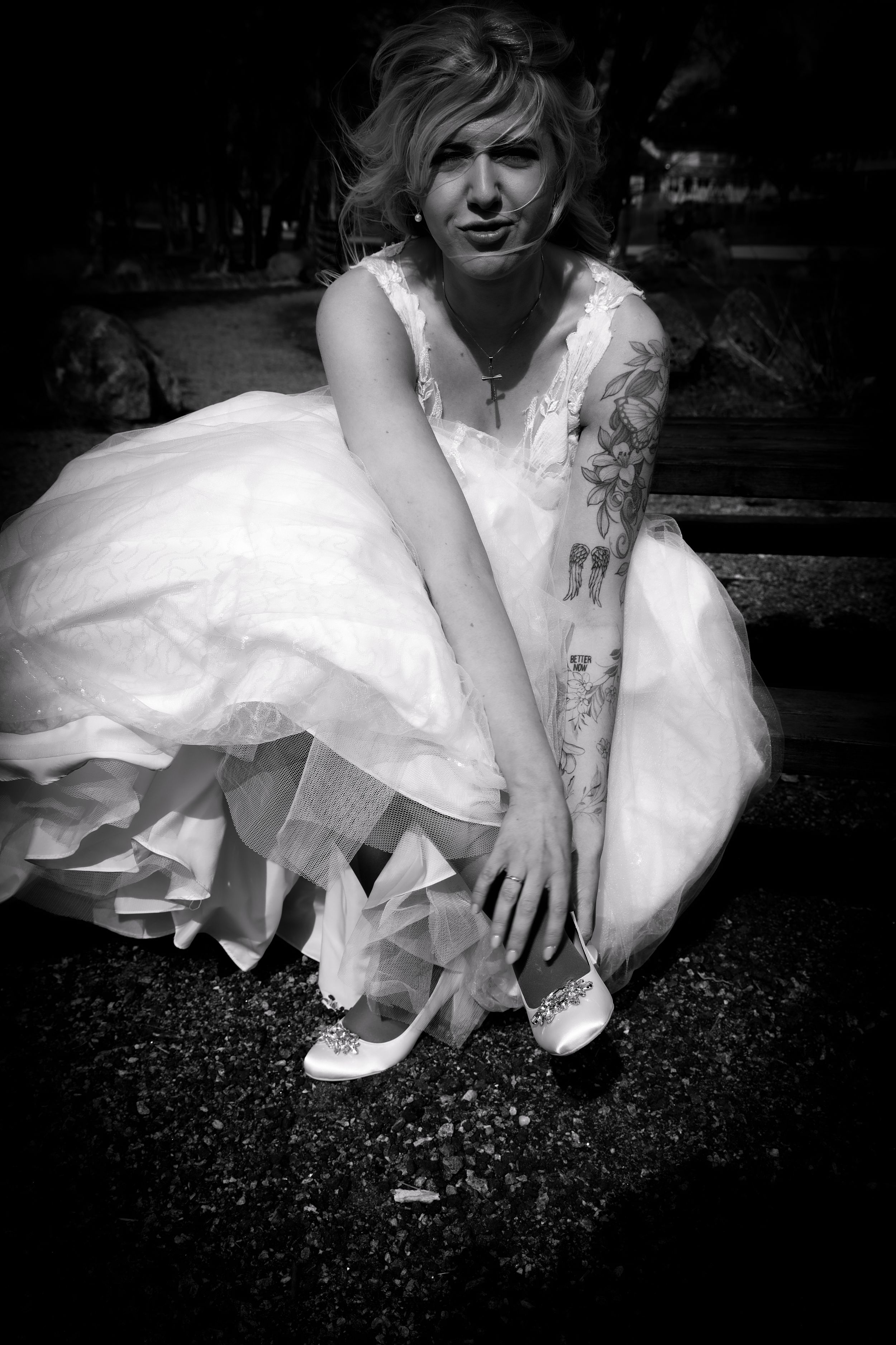 Black and white portrait of tattooed bride in white gown adjusting her heels outdoors.