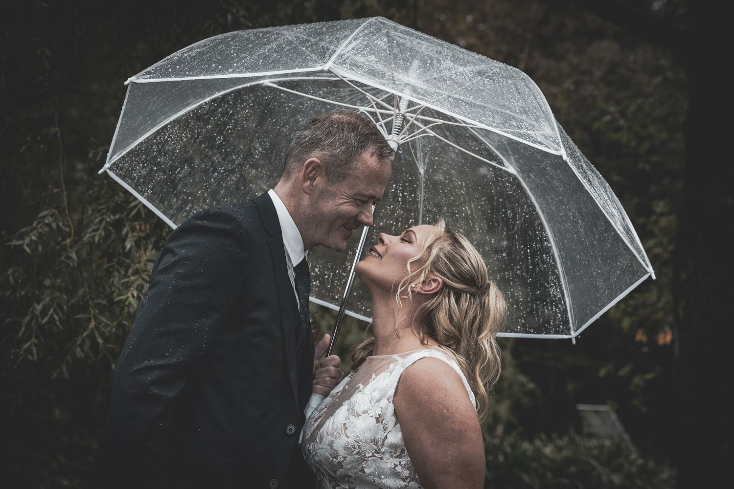 Wedding couple kissing tenderly under a transparent umbrella while rain falls around them.