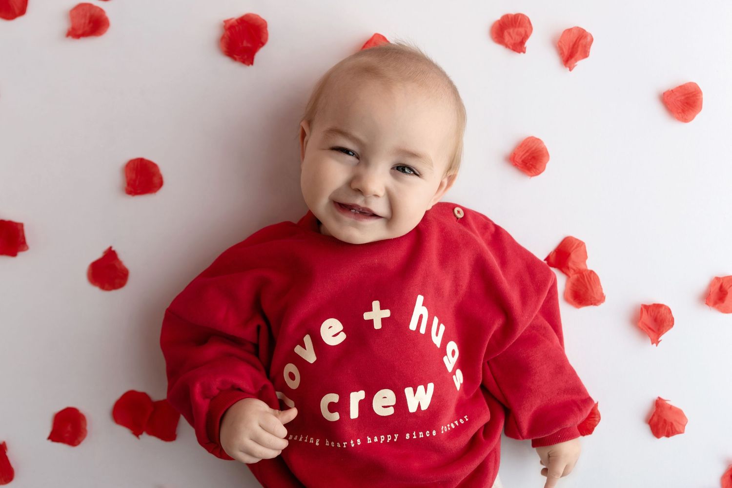 Smiling baby wearing red sweatshirt lying on white background with scattered red heart decorations.