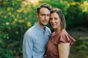 A couple embraces in a romantic outdoor photoshoot with golden sunlight filtering through green trees in the background.