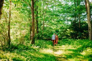 Two people walking together on a sunlit forest trail surrounded by lush green trees and vegetation.