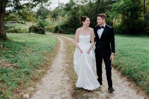 A couple in wedding attire walks along a winding dirt path surrounded by lush green grass and trees.