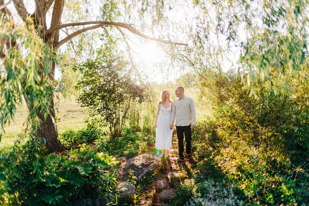 A group of people stand together under lush green trees in a garden setting during golden hour lighting.